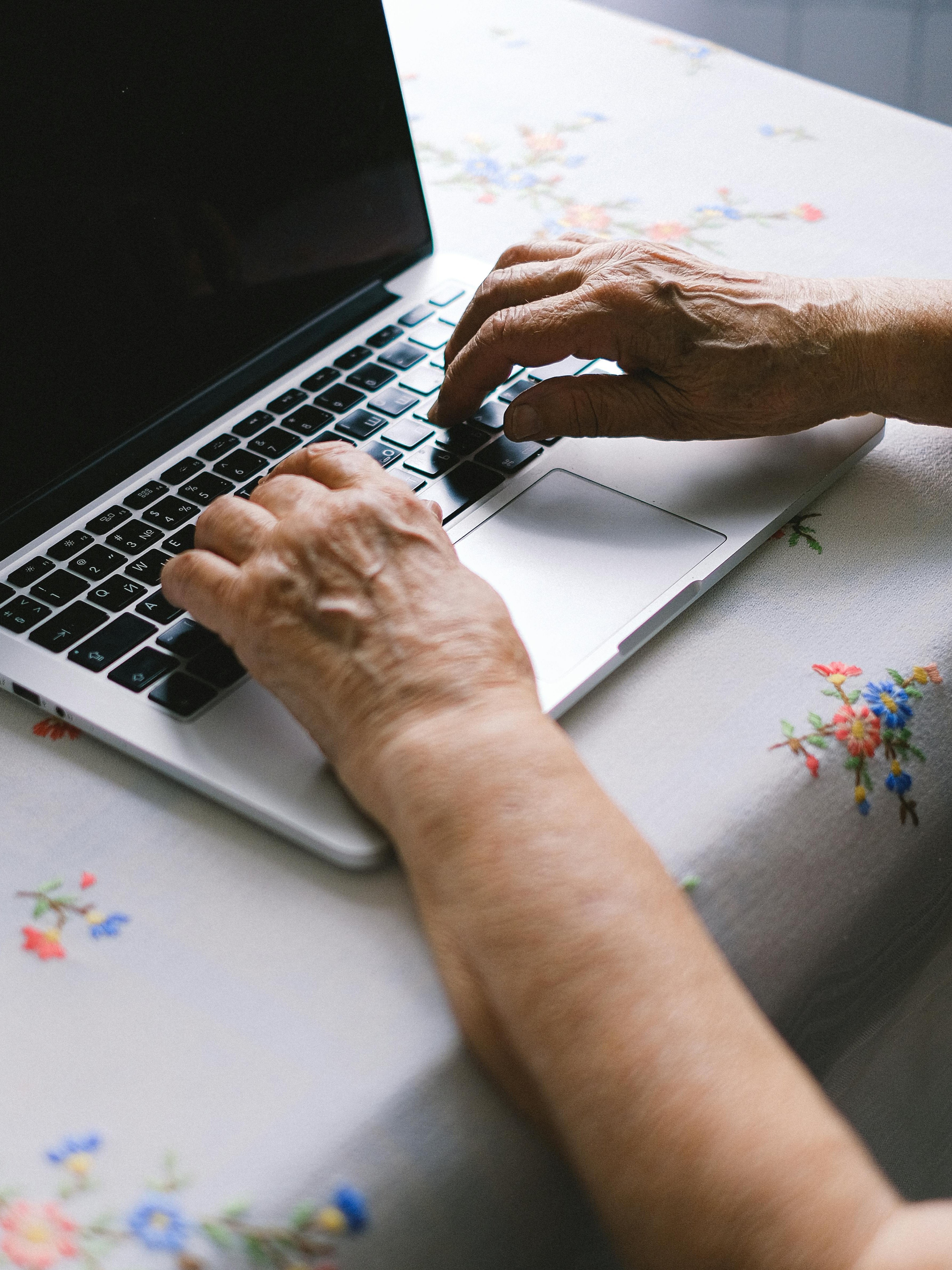 Technologie und Senioren im Homeoffice Hände einer älteren Person tippen auf einer Laptop-Tastatur auf einem blumigen Tischdecken.