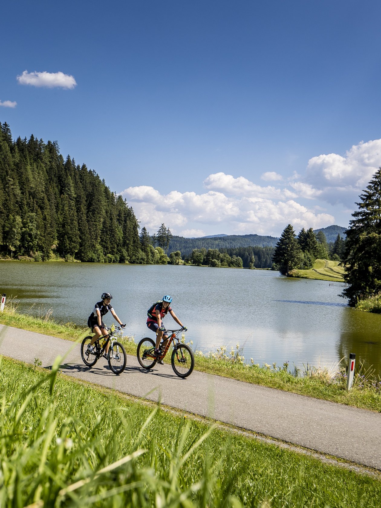 Mountainbike Abenteuer in der Natur Zwei Radfahrer auf einem Weg neben einem See, umgeben von Bäumen und Grünflächen.