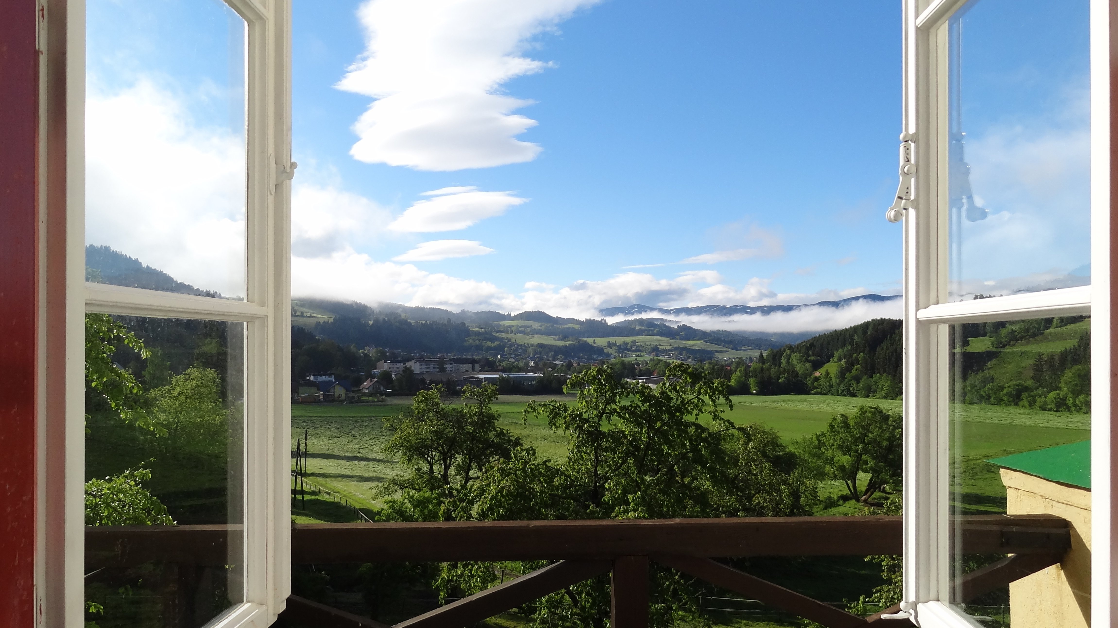 Offenes Fenster mit Bergblick Ein offenes Fenster mit Blick auf eine grüne Landschaft und Berge unter blauem Himmel
