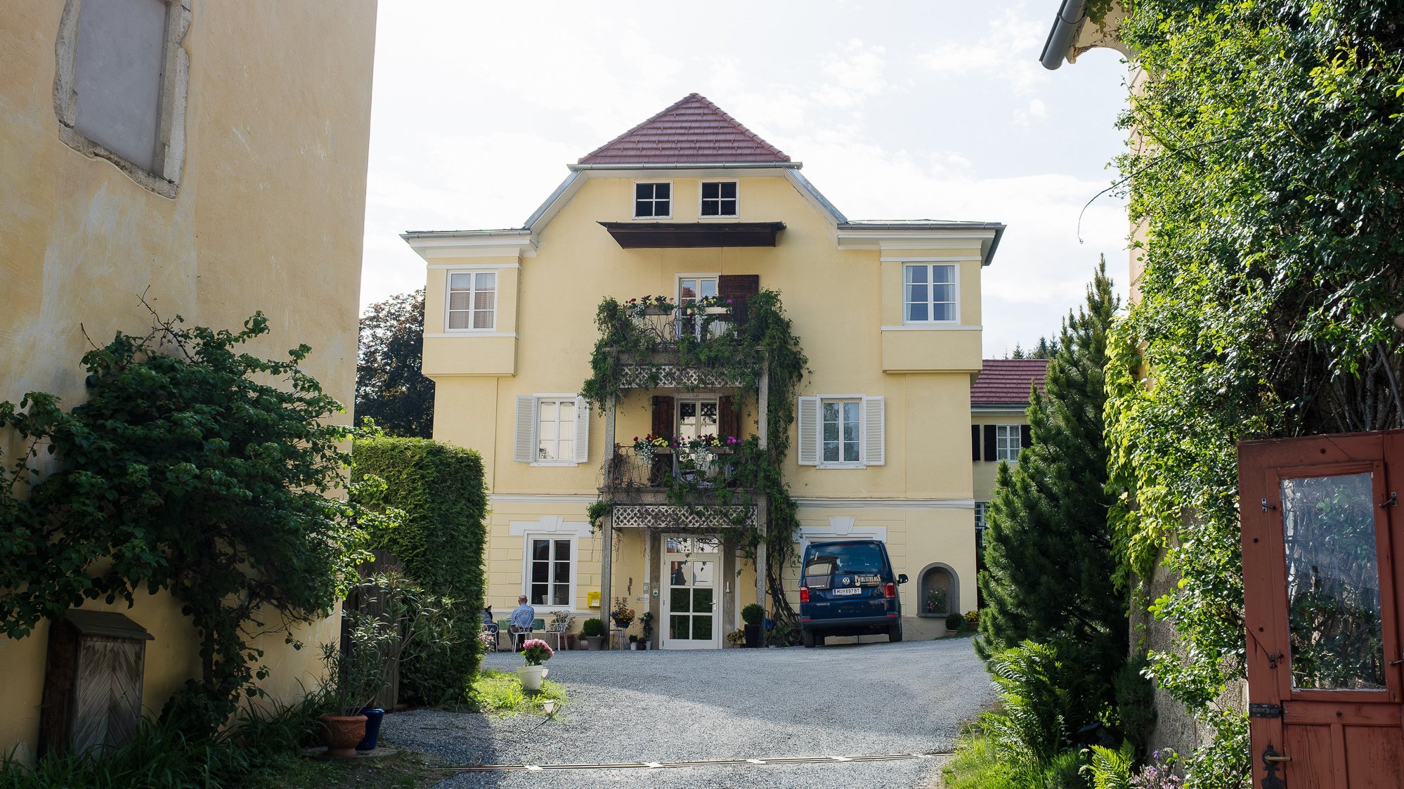 Zufahrt des Hotel Landsitz Pichlschloss Gelbes Wohngebäude mit Balkon und Pflanzen, umgeben von Garten und einer Kiesauffahrt.