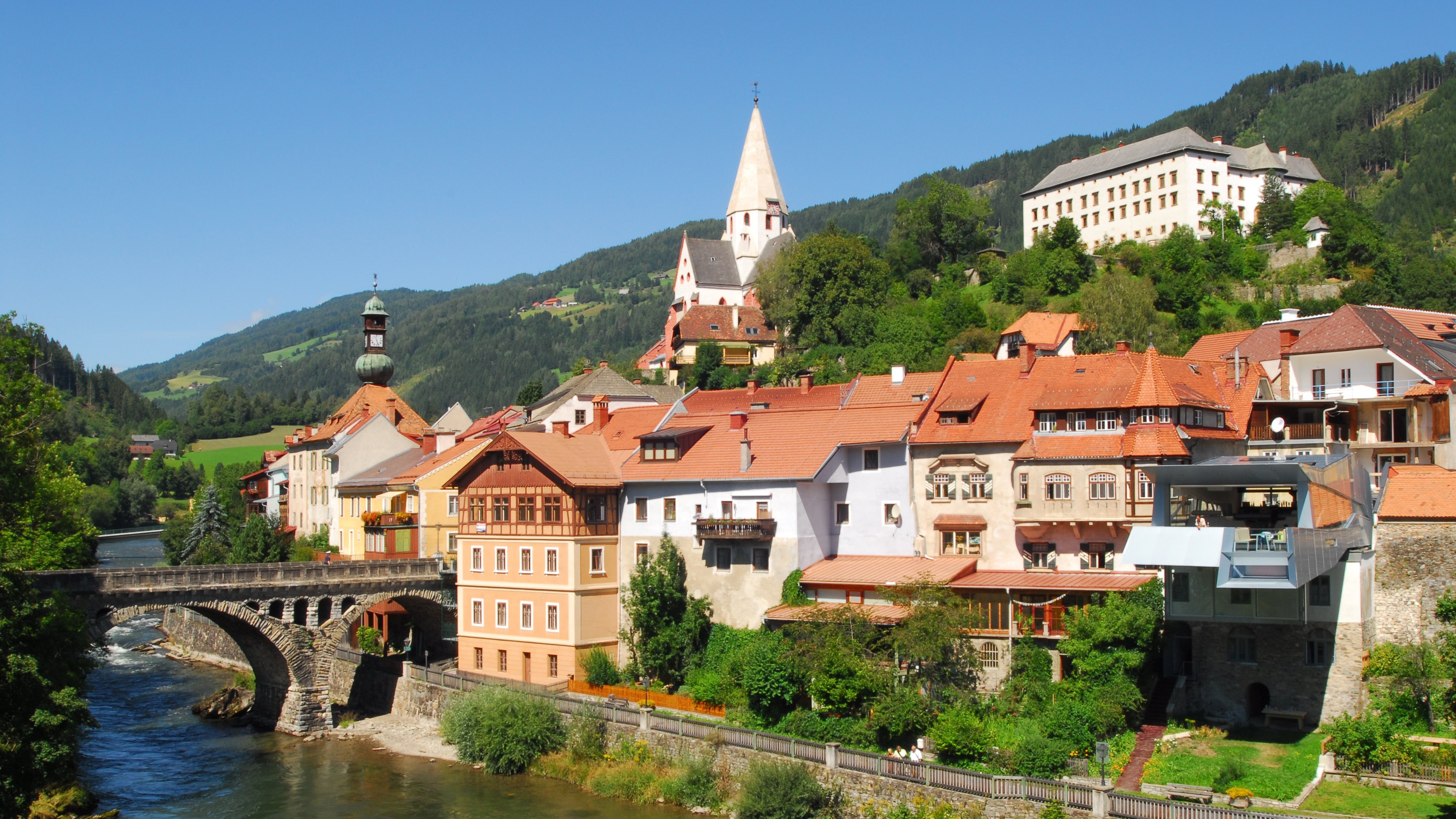 Malersches Dorf am Fluss Ein charmantes Dorf am Ufer eines Flusses mit historischen Gebäuden und Bergen im Hintergrund.