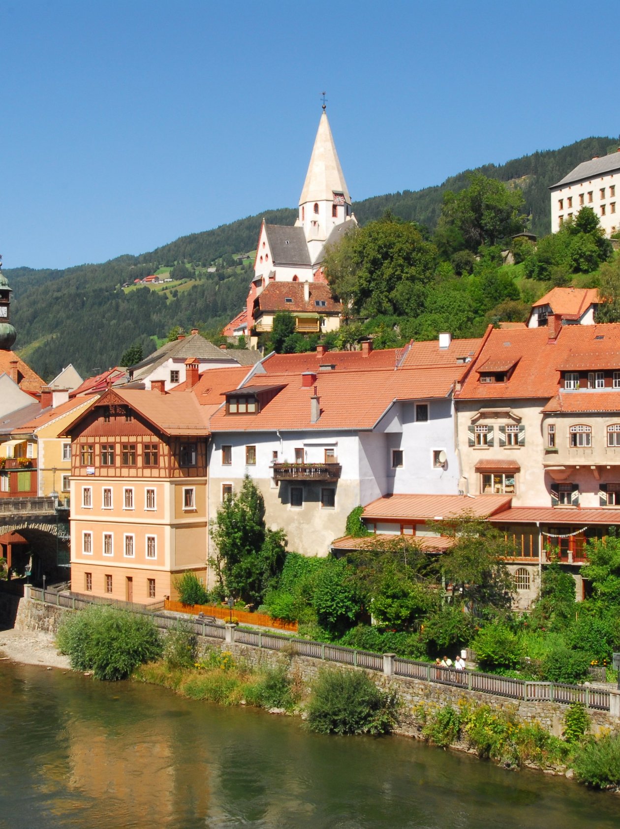 Malersches Dorf am Fluss Ein charmantes Dorf am Ufer eines Flusses mit historischen Gebäuden und Bergen im Hintergrund.