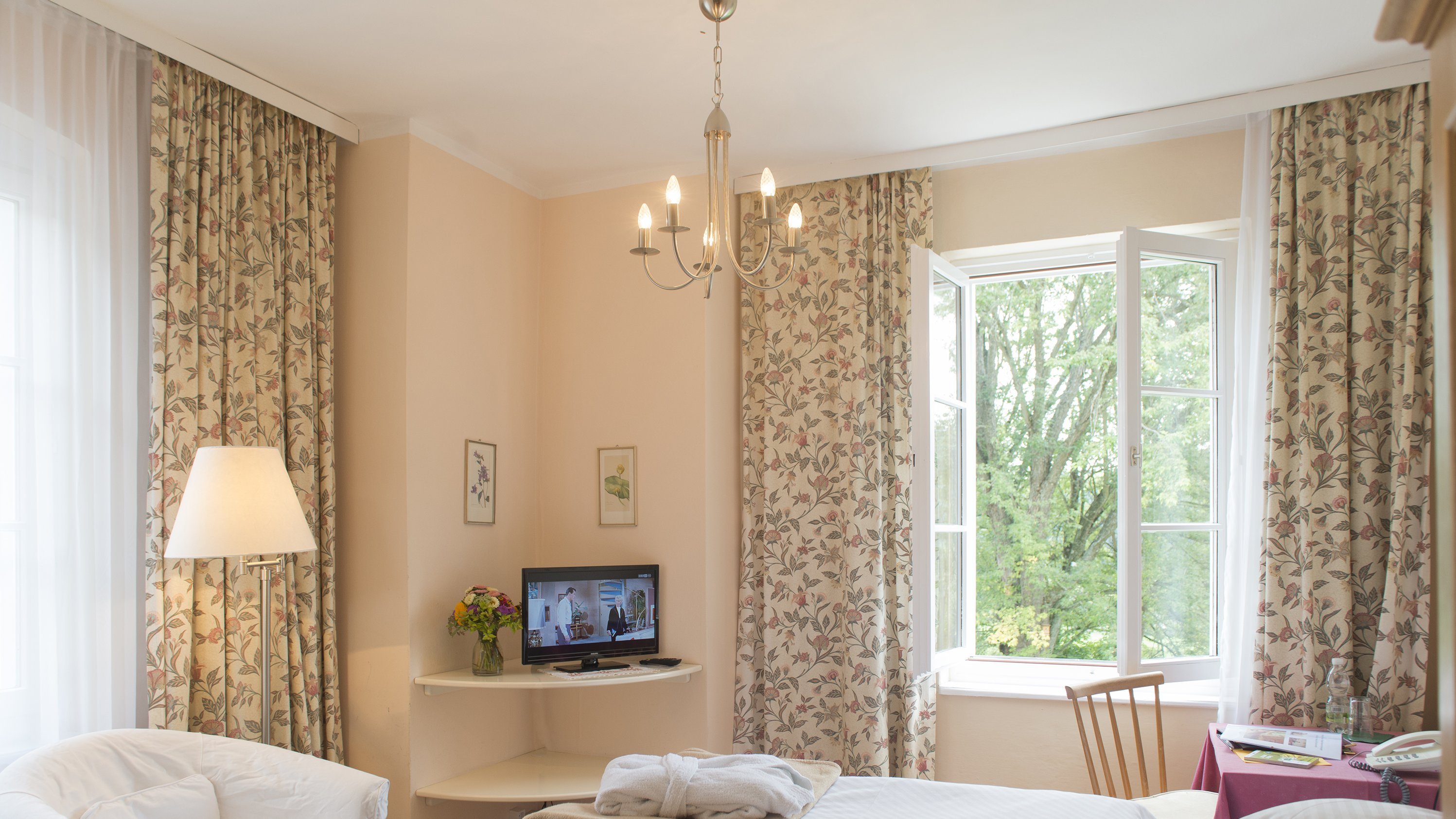 Bright and Cozy Hotel Room A well-lit hotel room with a bed, chair, television, and large windows adorned with floral curtains